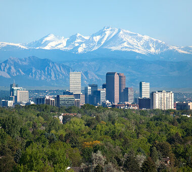 Snow covered Longs Peak, part of the Rocky Mountains stands tall in the background with green trees and the Downtown Denver skyscrapers as well as hotels, office buildings and apartment buildings filling the skyline.